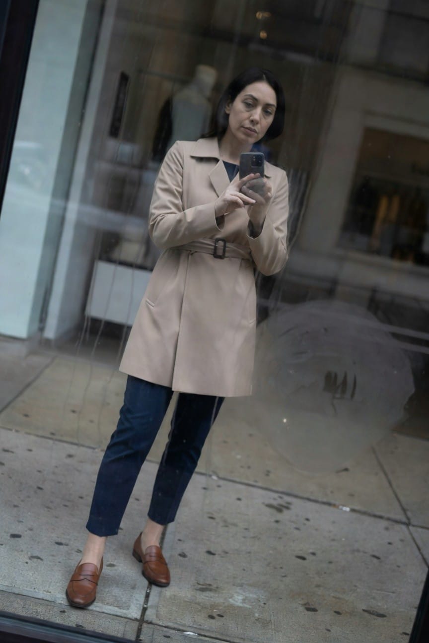 Woman standing outside a store window in a belted beige trench coat over slim blue trousers and brown leather loafers, taking a selfie with her phone