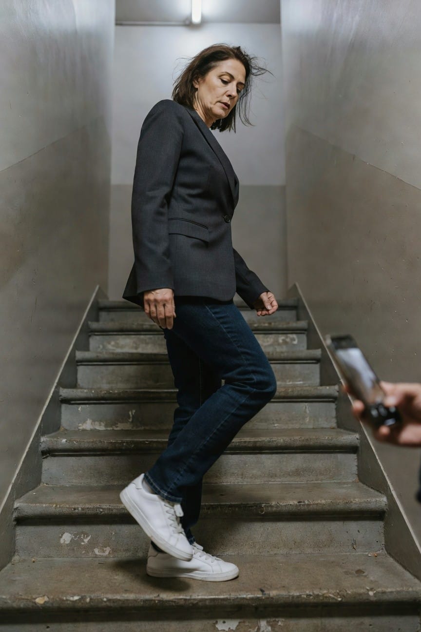Woman in her 50s wearing a dark gray tailored blazer over blue straight-leg jeans and white sneakers, standing dynamically on concrete stairs in a dimly lit stairwell, hand with phone visible in foreground