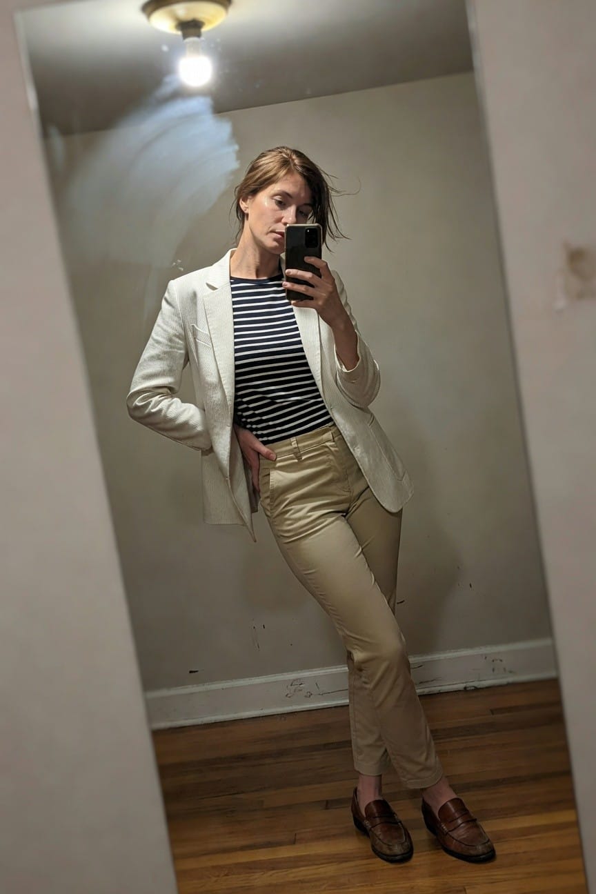 Mirror selfie of a woman in a cream textured blazer over a navy and white striped shirt, khaki satin pants, and brown loafers, posing casually in a simply lit room