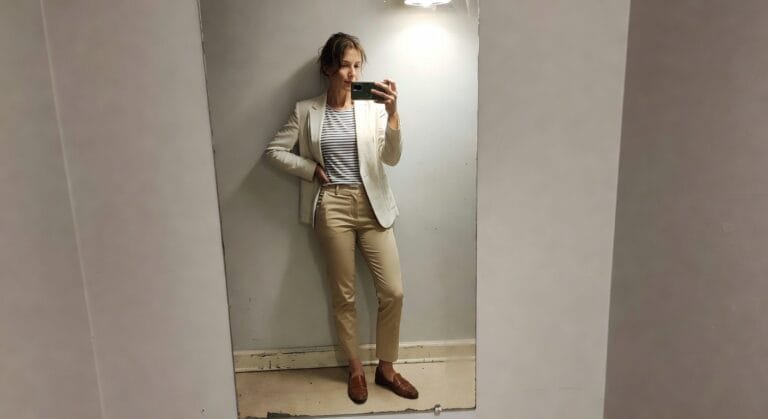 Mirror selfie of a woman in a cream textured blazer over a navy and white striped shirt, khaki satin pants, and brown loafers, posing casually in a simply lit room