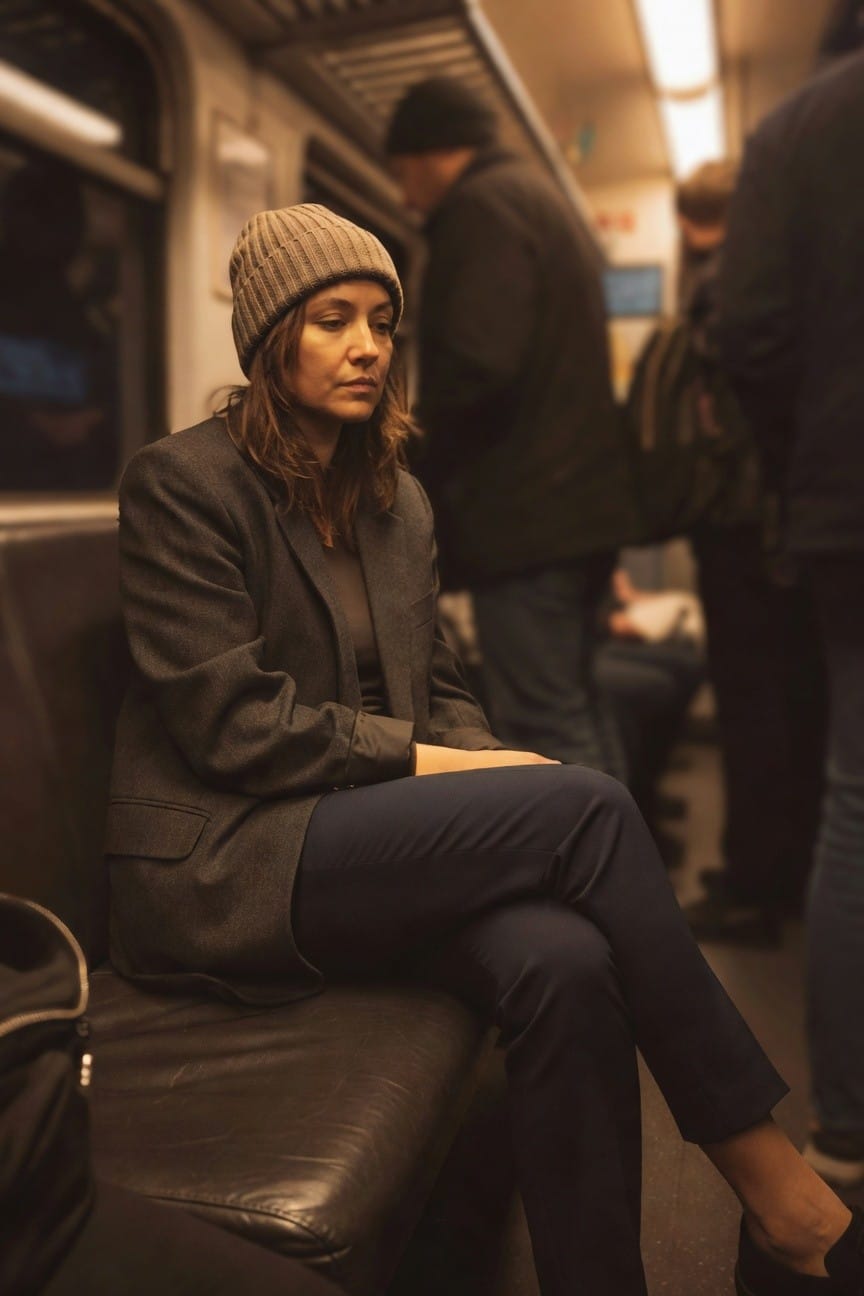 Woman seated wearing a beige ribbed knit beanie, gray tweed blazer over a dark top, slim navy trousers, black loafers, and a black leather bag beside her on a leather bench