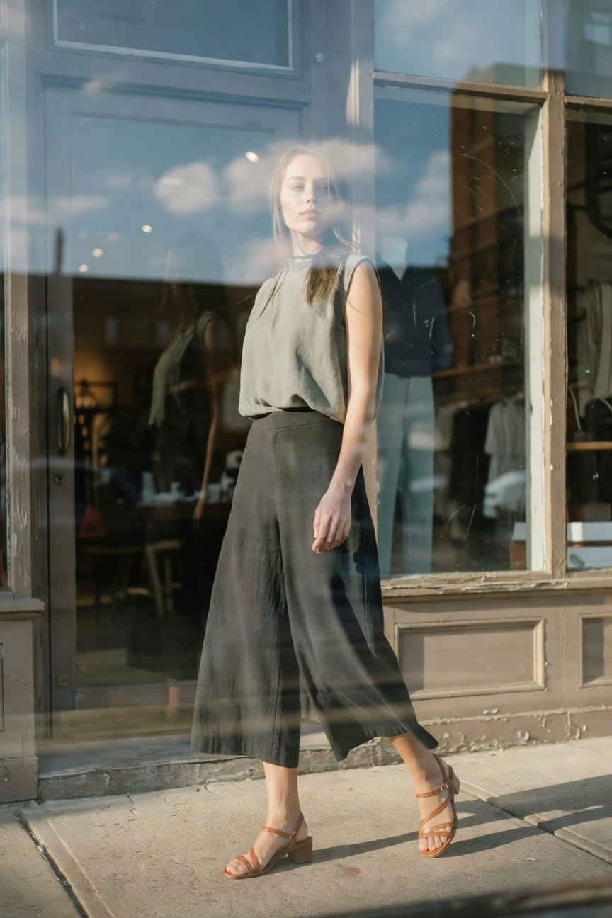 Woman outside a storefront window in a sleeveless sage green halter top, black wide-leg linen pants, tan strappy heeled sandals, and a small black crossbody bag, posing casually with one hand in pocket