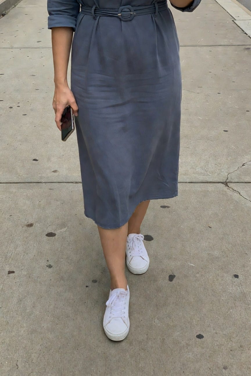 Side view of a woman wearing a belted dusky blue-gray midi dress with rolled sleeves, holding a phone, paired with white low-top sneakers on a sidewalk