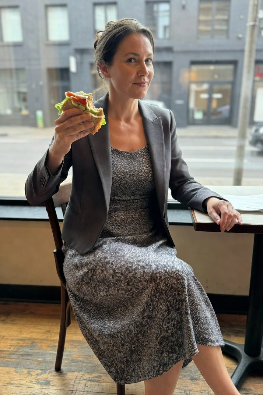 Woman in gray blazer over gray patterned midi dress, seated at cafe table holding sandwich, urban window view behind