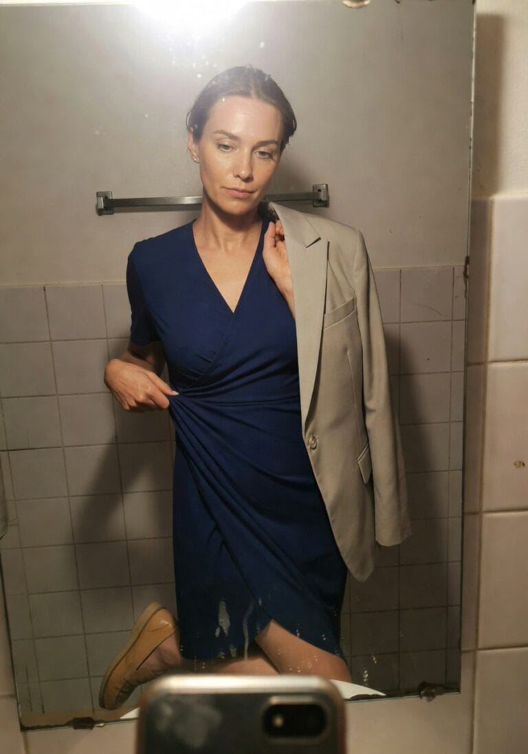 Woman taking a mirror selfie in a bathroom wearing a navy blue wrap dress with a gray blazer draped over one shoulder, holding her phone, brown loafers on feet, towel hanging nearby