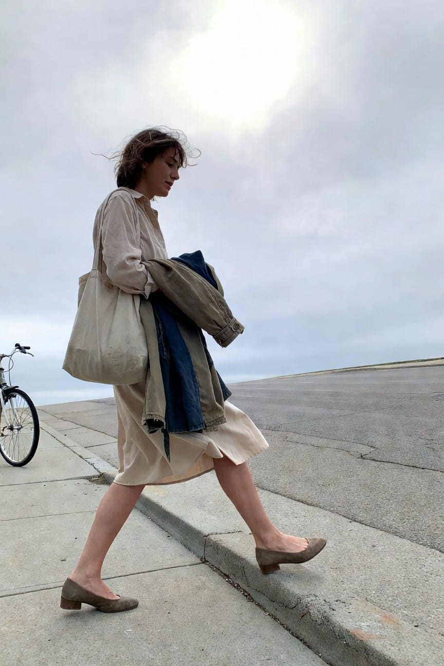 Woman walking in a loose cream long-sleeve shirt dress with a navy jacket draped over her arm and skirt, beige canvas tote bag on shoulder, and brown suede low-heeled pumps, casual work-appropriate styling on a concrete path