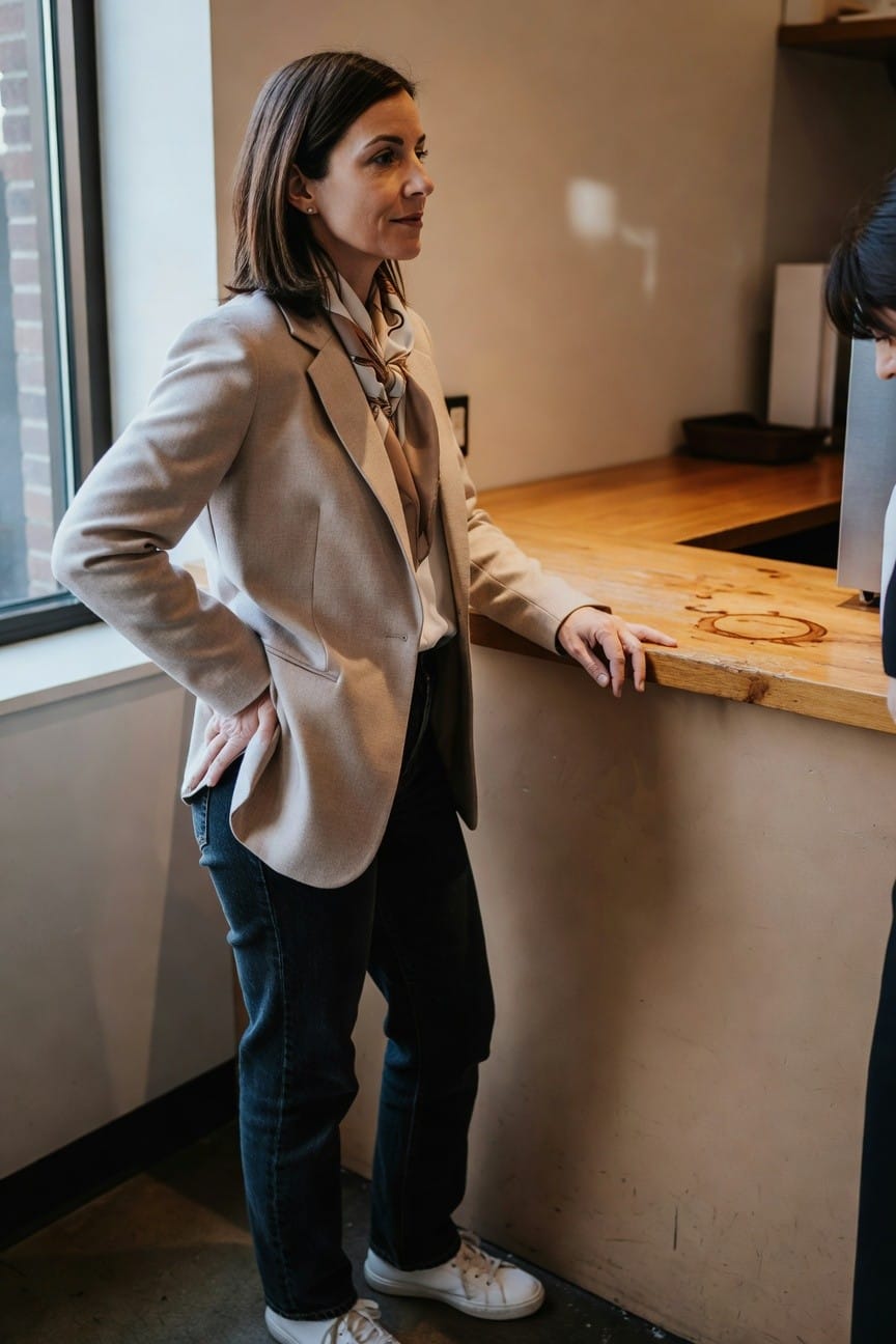 Woman in beige blazer over white shirt with silk scarf, dark straight-leg jeans, and white sneakers, leaning casually on a wooden counter in a modern kitchen space