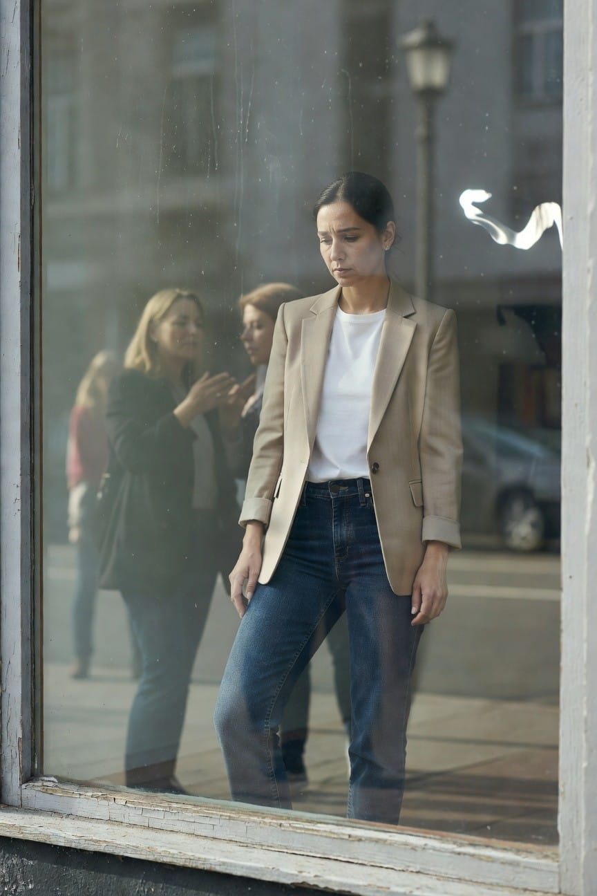 Woman reflected in a shop window wearing a beige blazer over white t-shirt and blue straight-leg jeans standing casually on a street with blurred background figures