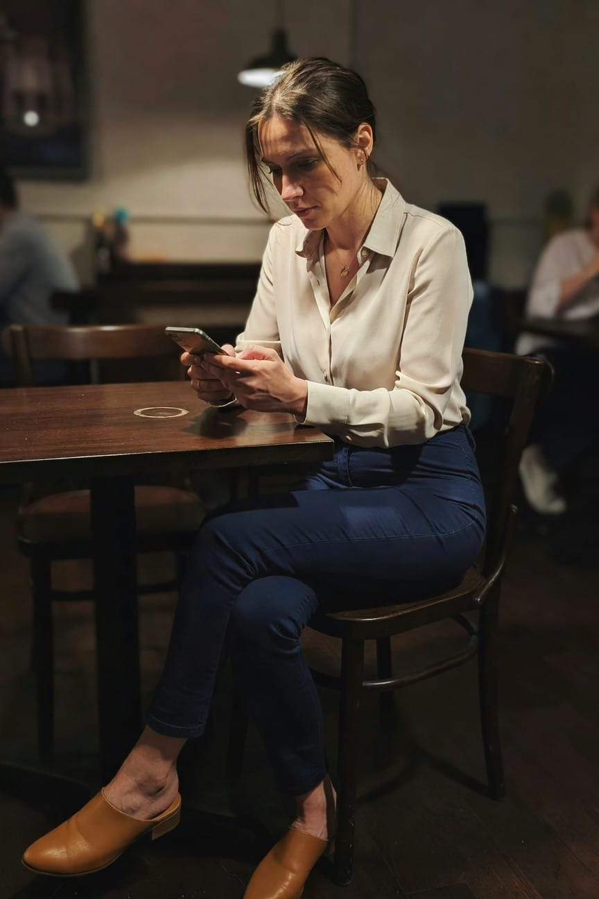 Woman in cream silk blouse, navy pants, and tan wooden clogs sitting at a wooden table while checking her phone in a dimly lit cafe