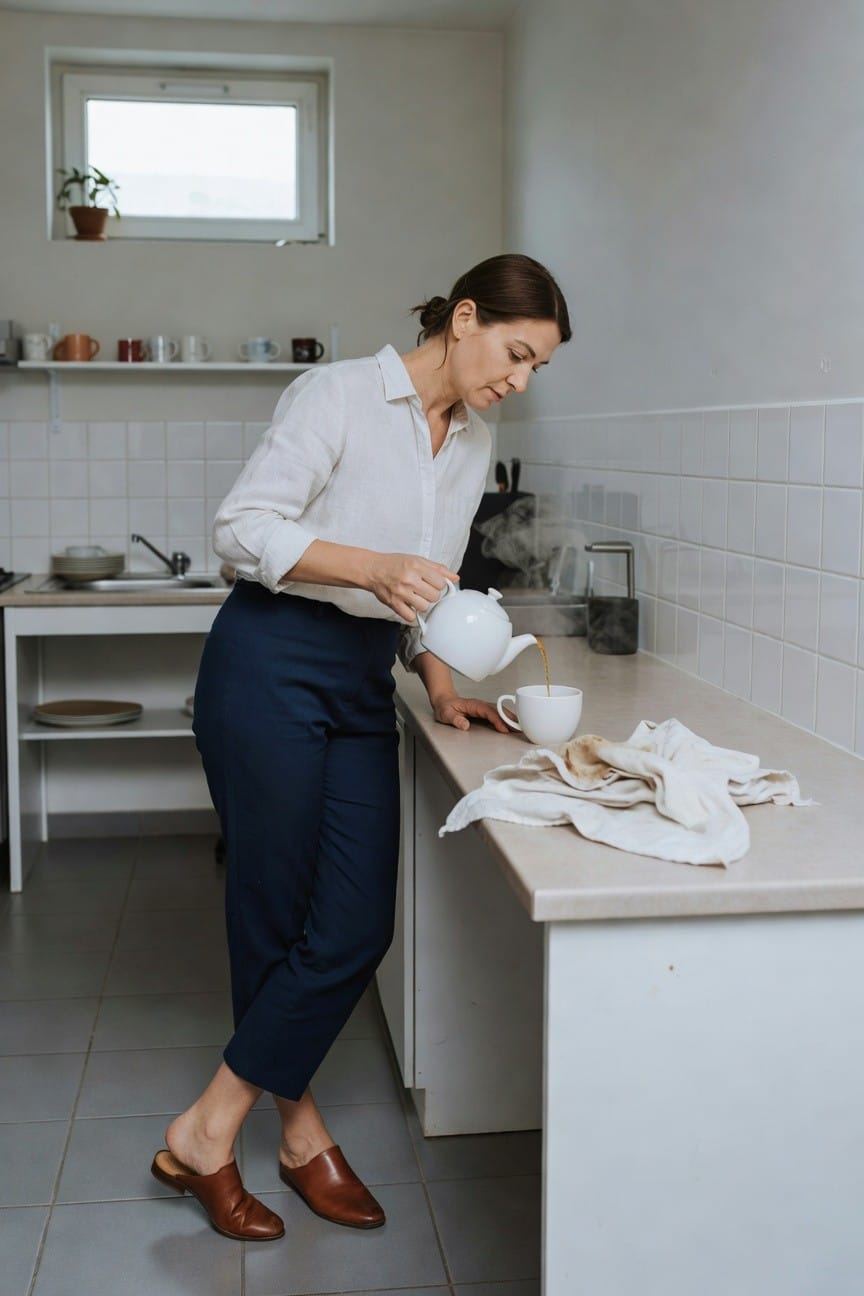 Woman in white linen shirt, navy cropped trousers, and brown leather mules standing in a kitchen while pouring tea from a white teapot into a cup
