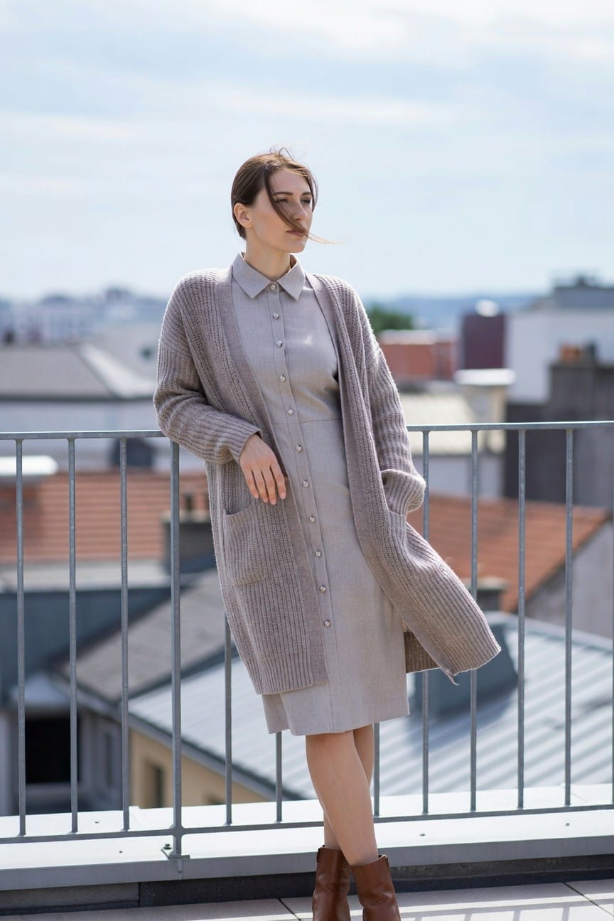 Woman in light gray button-front shirt dress layered with open long gray knit cardigan, leaning on metal balcony railing, wearing brown ankle boots, urban rooftop view in background