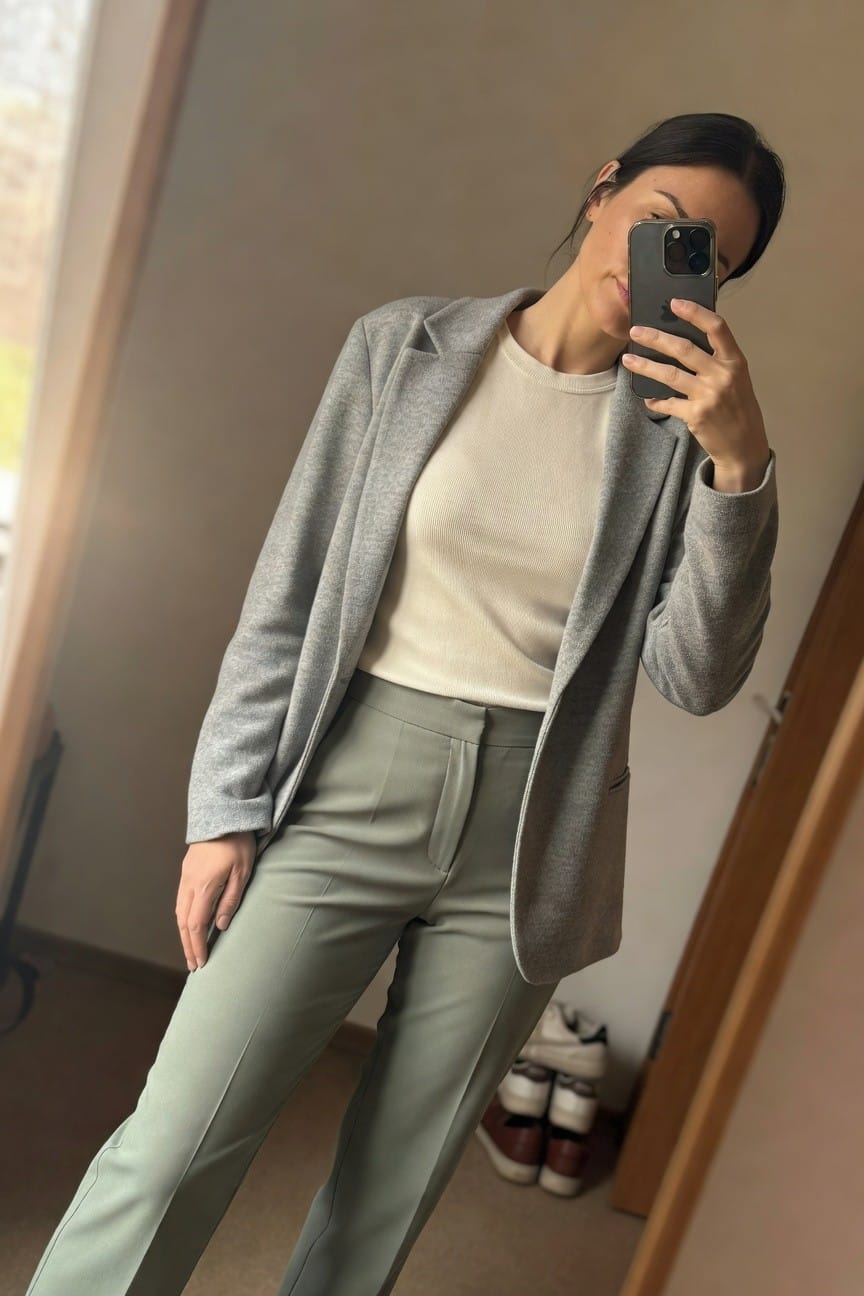 Woman taking a mirror selfie in an open gray knit blazer over cream turtleneck top and high-waisted sage green tailored trousers standing in a simply furnished room with wooden doors and sneakers on the floor