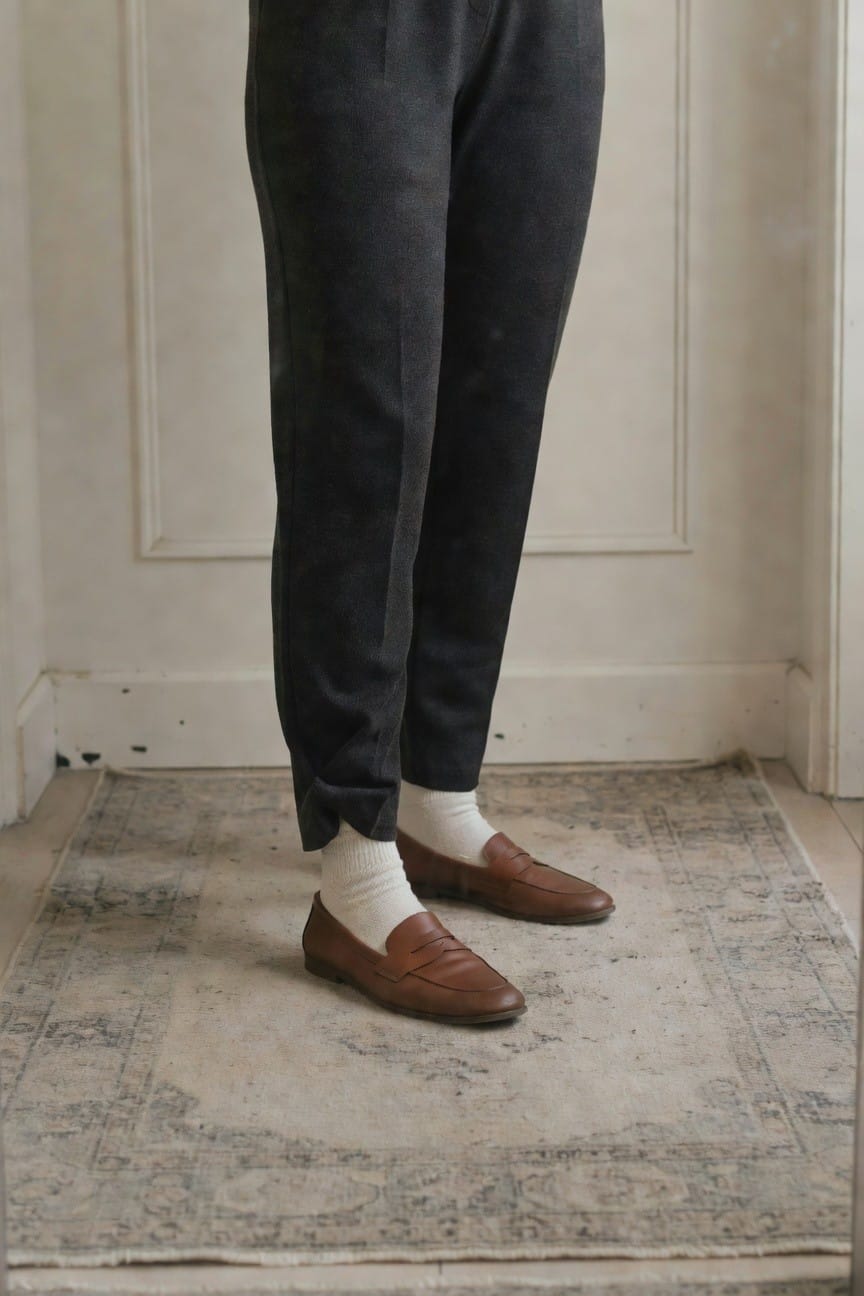 Woman's lower legs in dark gray pleated trousers cuffed at ankles revealing white socks and brown woven loafers on a patterned rug against white paneled wall