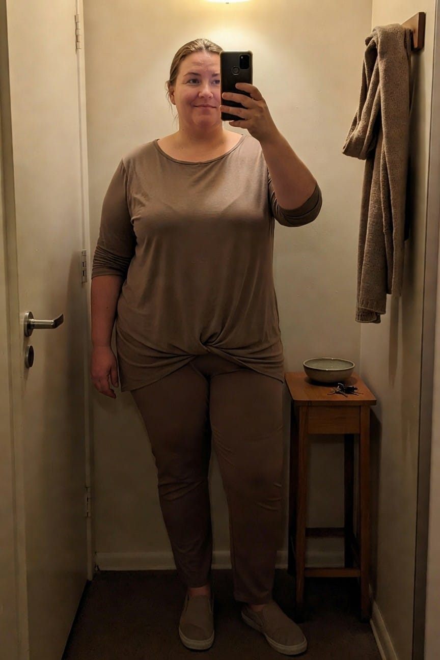 Plus-size woman smiling in a selfie, wearing a loose taupe twist-front top with three-quarter sleeves, matching wide-leg taupe pants, and beige slip-on sneakers, standing in a small bathroom.