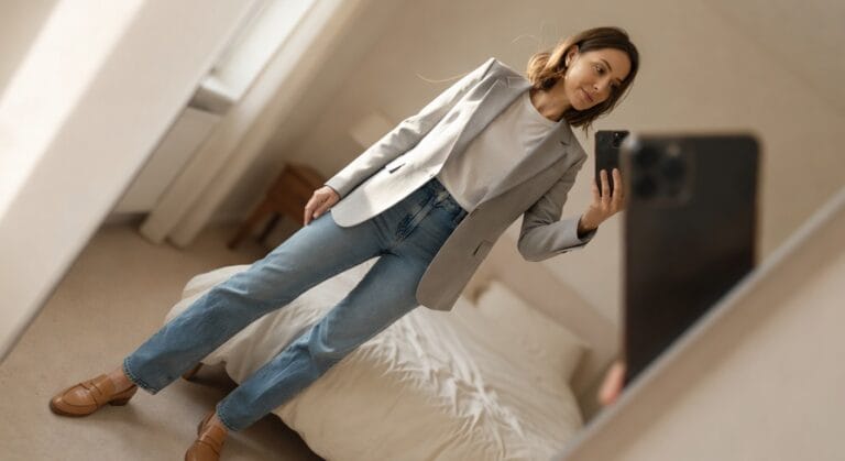 Woman taking a mirror selfie in a light gray tailored blazer over white t-shirt, high-waisted light blue straight-leg jeans, brown leather loafers, minimal jewelry, neutral bedroom background