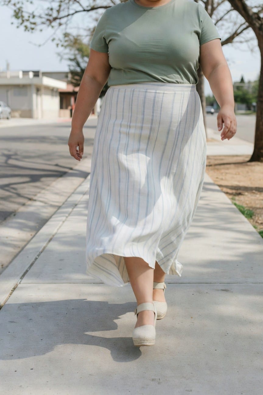 Plus-size woman walking on sidewalk in sage green short-sleeve t-shirt, white striped maxi skirt with side slit, and beige espadrille wedge sandals, side profile view