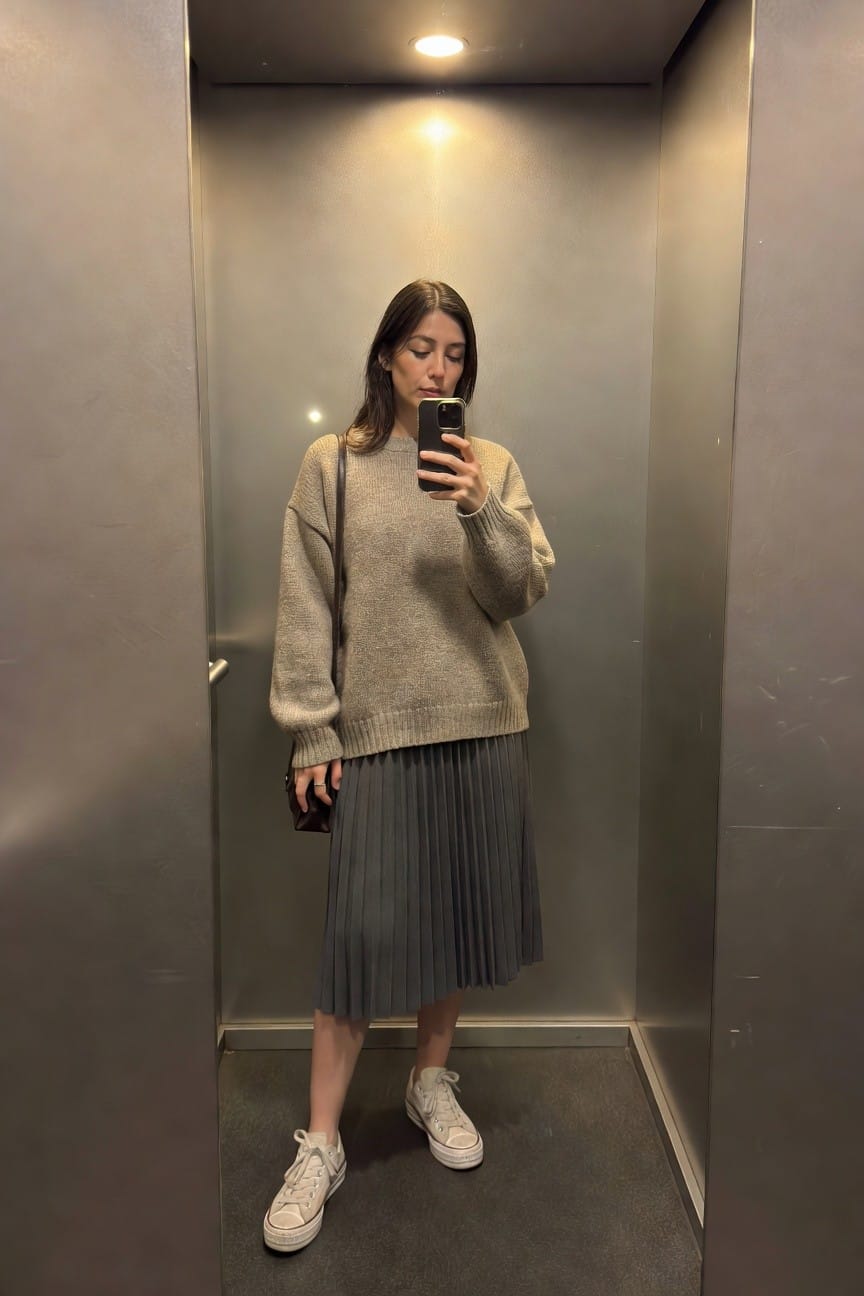 Woman taking a mirror selfie in an elevator wearing a loose beige knit sweater, dark gray pleated midi skirt, white low-top sneakers, and a small brown crossbody bag, with brunette hair loose over shoulders