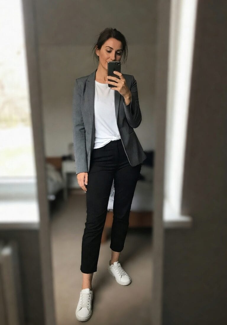 Woman taking mirror selfie in gray blazer over white t-shirt, black ankle-length pants, and white sneakers, hand in pocket, bedroom background