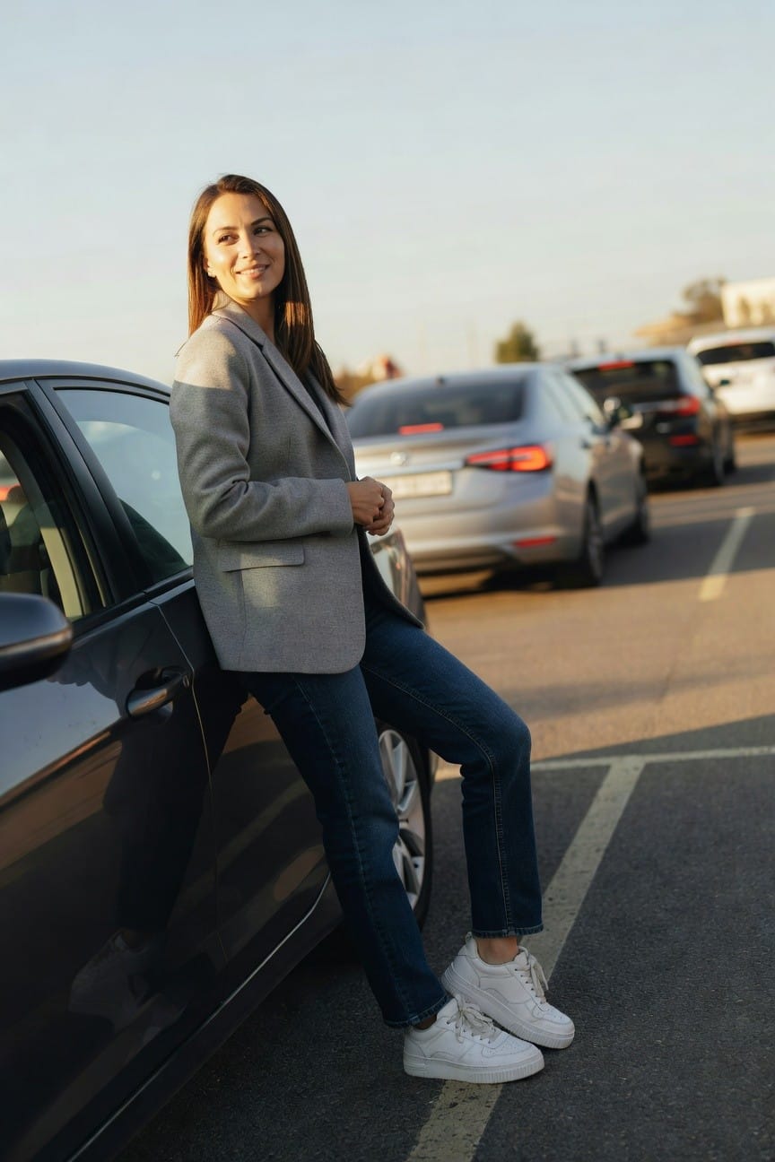 Woman in light gray tailored blazer over blue straight-leg jeans and white low-top sneakers, leaning casually against a car door