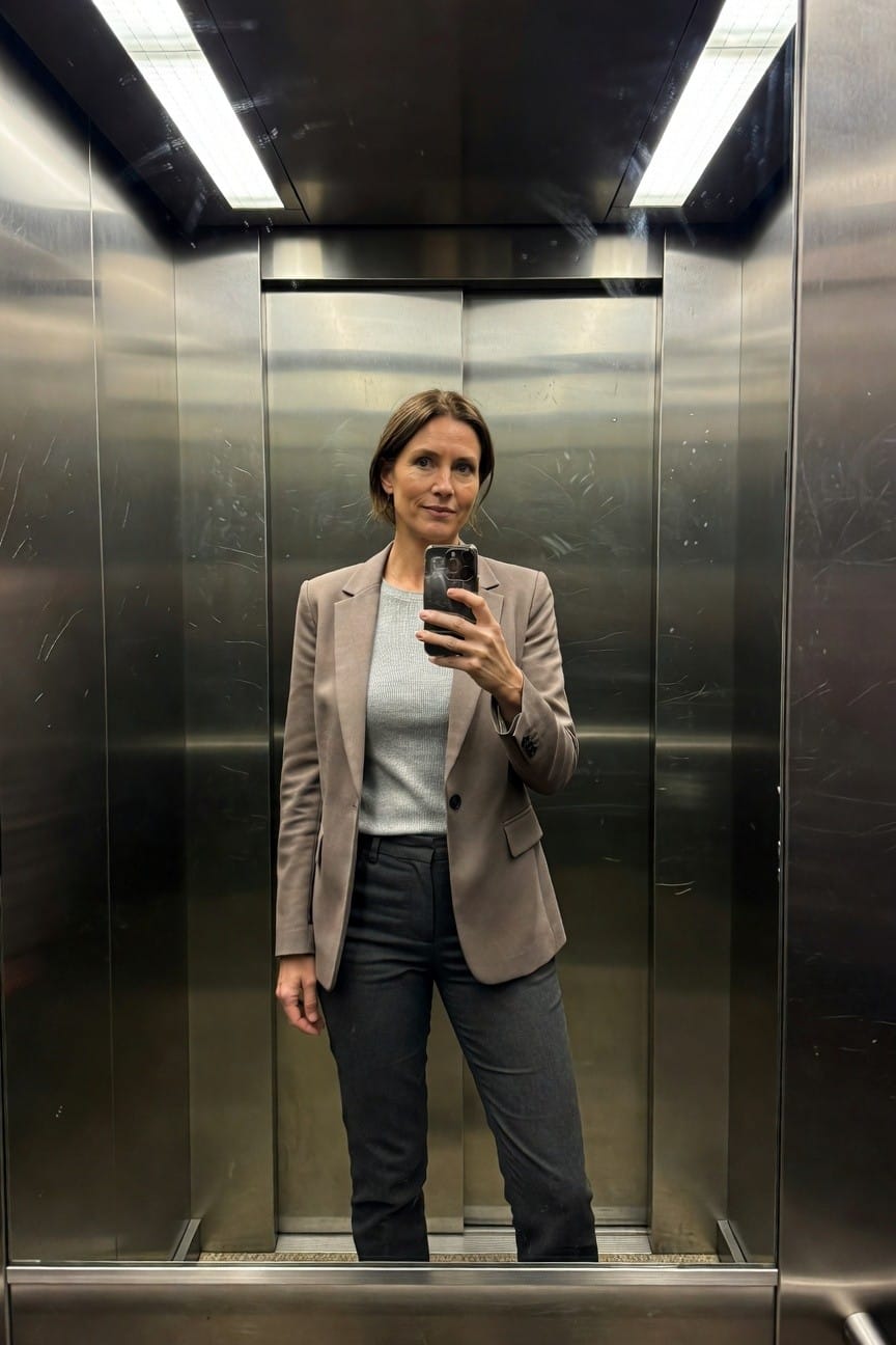 Woman in taupe single-breasted blazer over light gray knit sweater and dark gray straight-leg trousers, posing in mirror selfie inside steel elevator with phone in hand