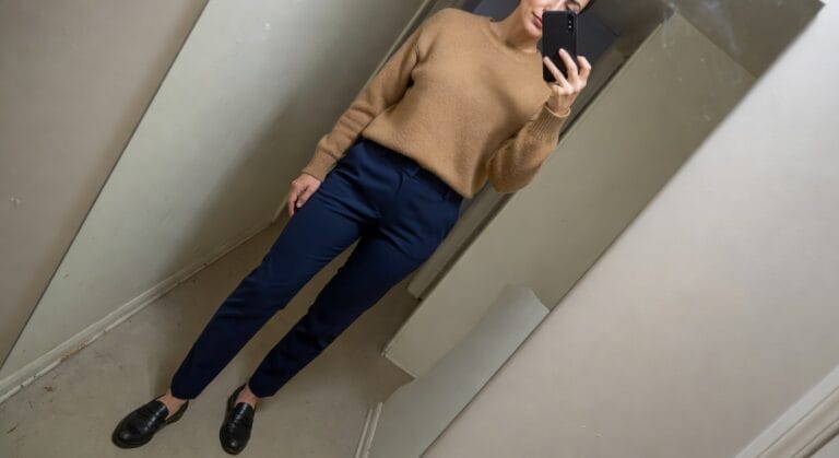 21 Comfortable Classy Work Outfits For Women Who Need All Day Style And Ease Woman taking mirror selfie in oversized camel knit crewneck sweater, navy pleated tailored trousers, and black leather loafers, standing confidently with hands on hips