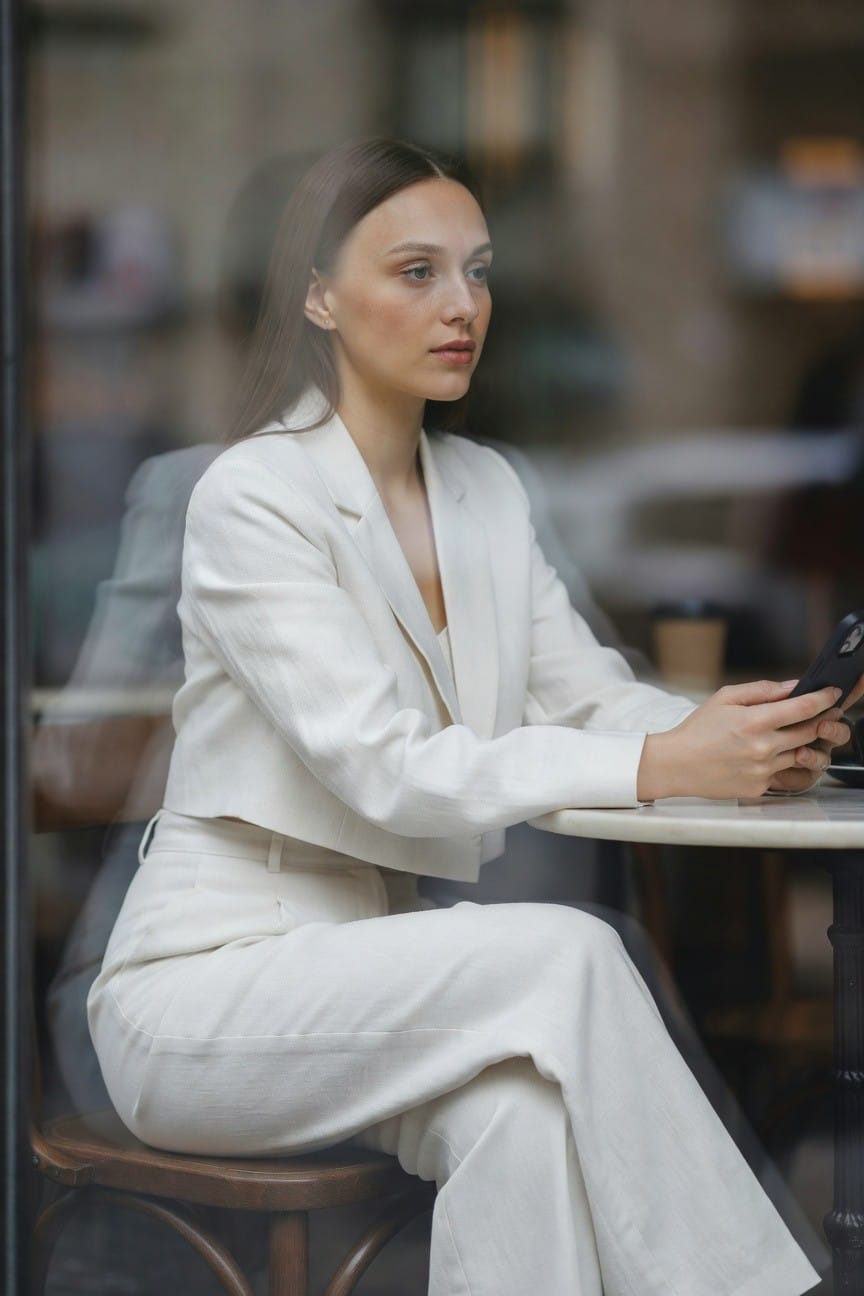 Woman in a white tailored cropped blazer and matching high-waisted wide-leg pants sits at a cafe table holding a phone, elegant professional outfit with subtle fabric texture