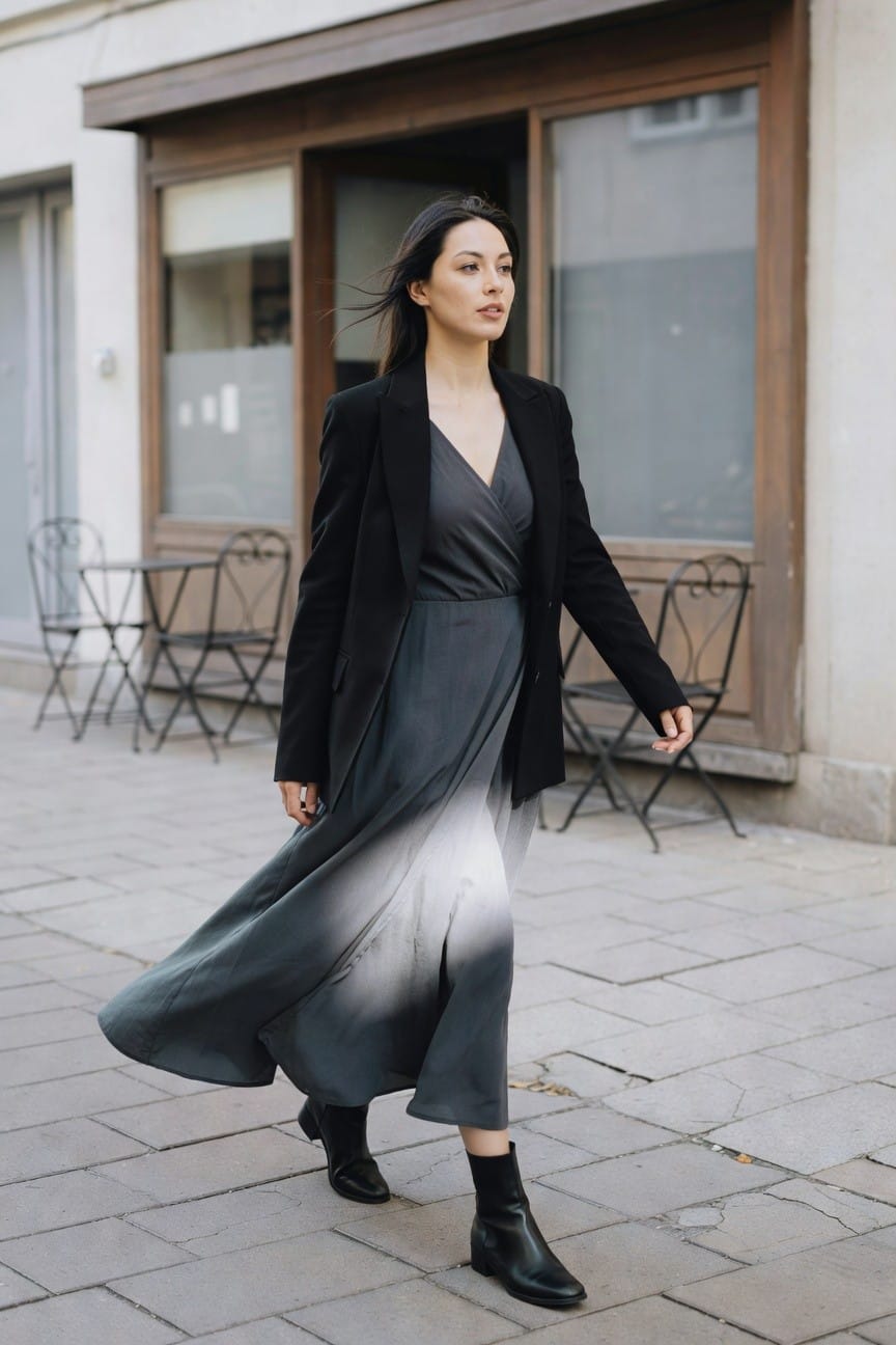 Woman in open black blazer over gray-to-white ombre wrap dress with black ankle boots, walking on paved street outside European-style cafe