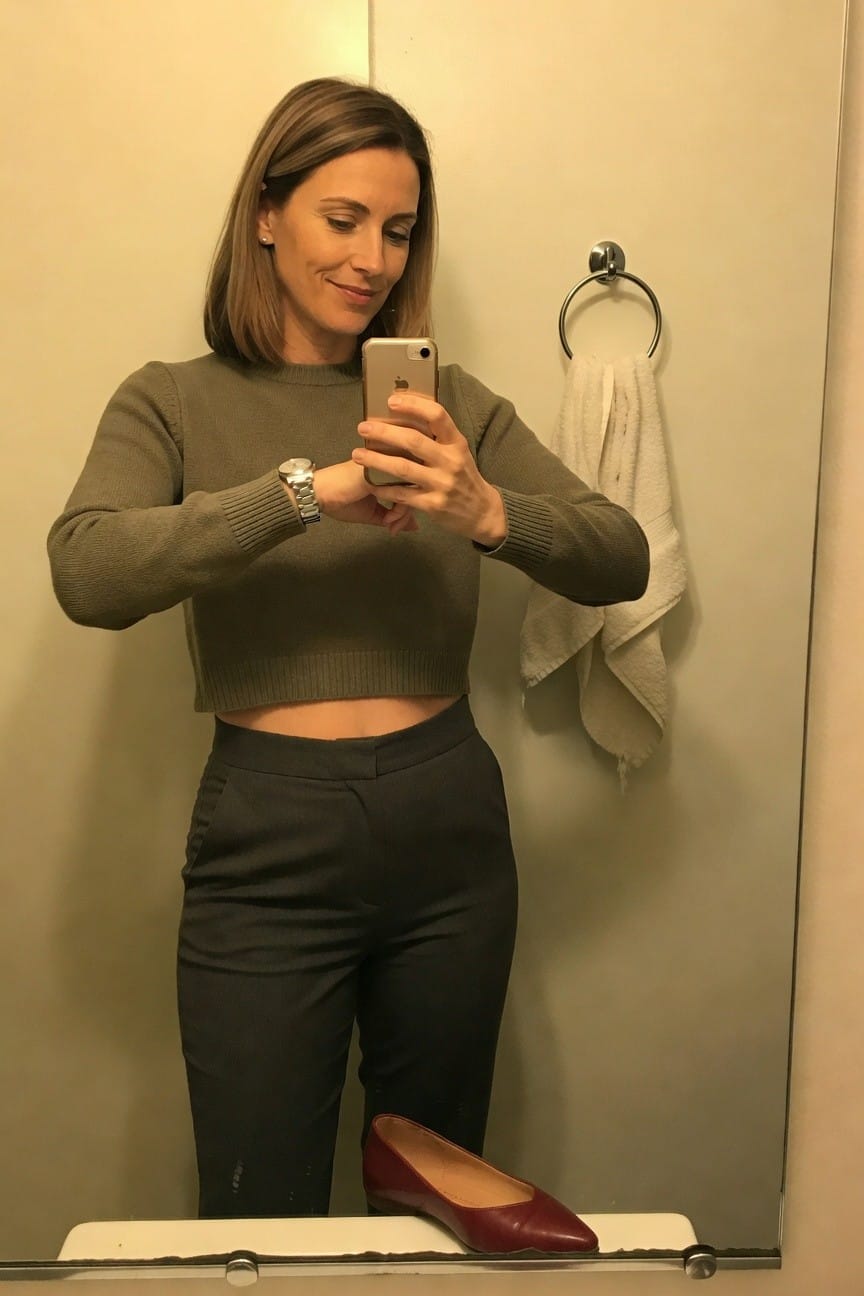 Woman in mirror selfie wearing long-sleeve cropped olive green knit sweater showing midriff, high-waisted charcoal gray tailored trousers, silver watch on wrist, red pointed-toe heels on bathroom floor below