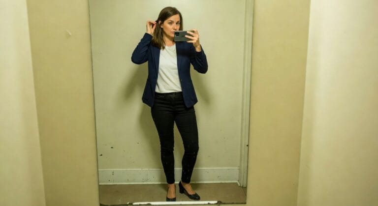 Woman taking a mirror selfie in a navy pinstripe blazer over white t-shirt, black slim pants, and black block-heel pumps, adjusting her bob haircut in a fitting room