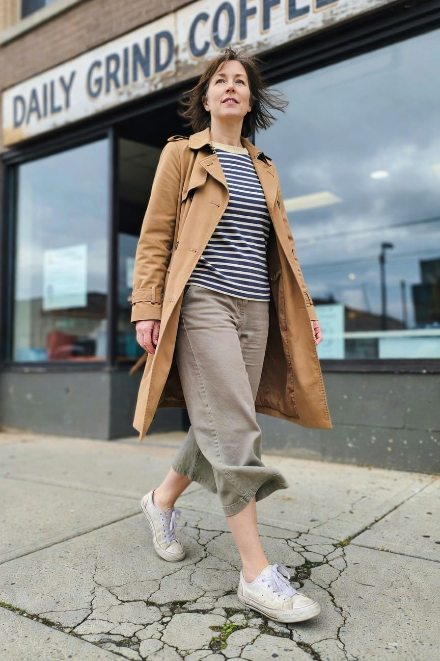 Woman in her 40s with shoulder-length brown hair wearing an open beige trench coat over a navy-and-white striped Breton shirt, gray cropped wide-leg pants, and white canvas sneakers, standing on a cracked sidewalk in front of a Daily Grind Coffee shop window