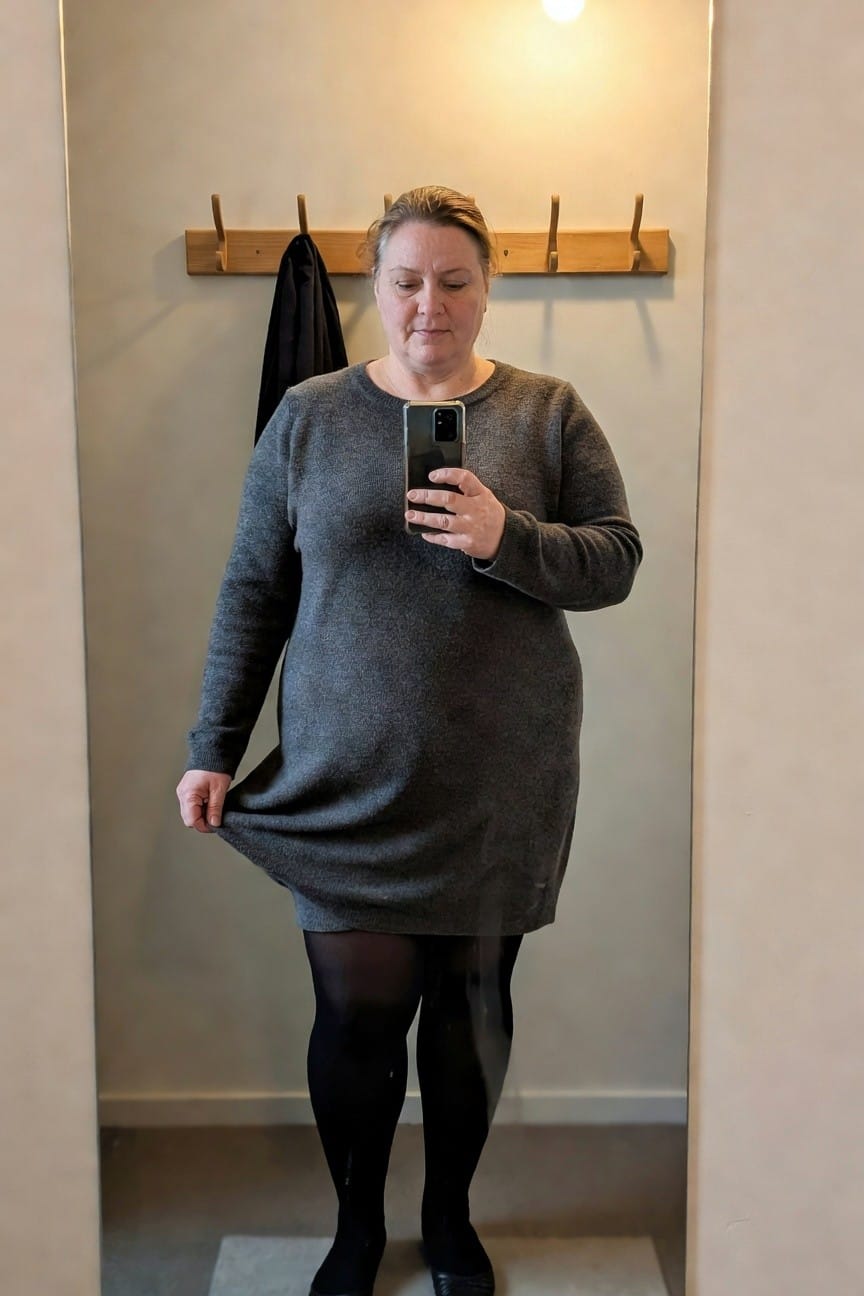 Plus-size woman taking a mirror selfie in a long-sleeved gray knit sweater dress with A-line skirt, black sheer tights, and black flats, standing in a hallway with coat rack nearby
