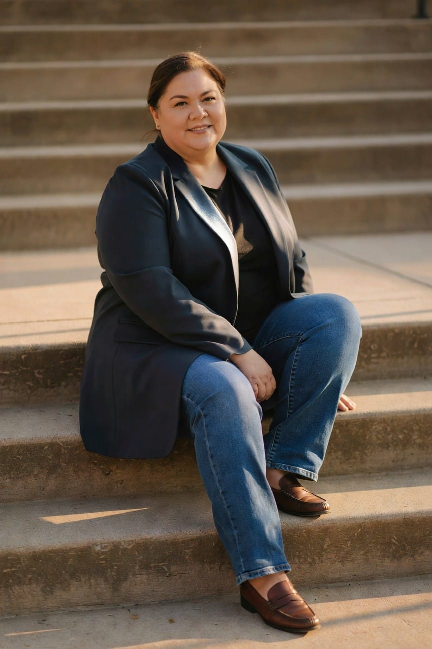 Plus-size woman seated wearing a long navy blazer over black top, blue jeans, and brown loafers, professional casual work outfit on concrete steps