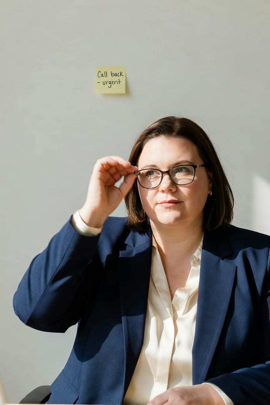 Plus-size woman with bob haircut and glasses adjusting her eyewear while seated, wearing a navy blue blazer over a white blouse with pearl earrings and bracelet in an office setting