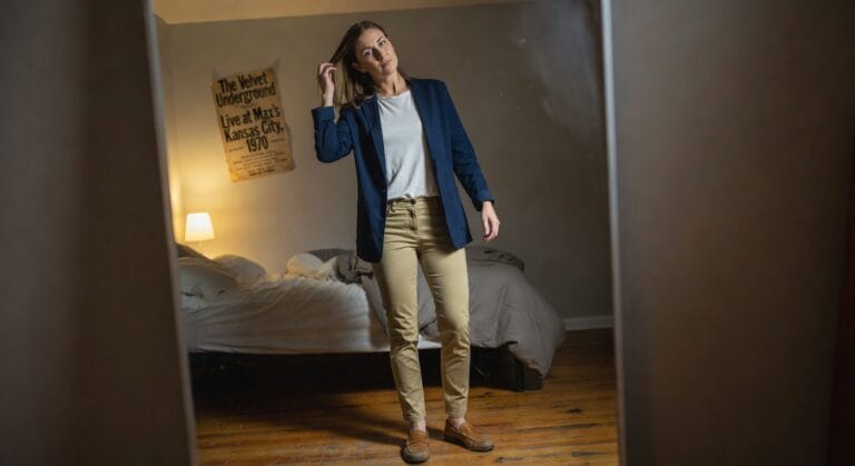 Woman taking a mirror selfie in a navy blazer over white t-shirt, khaki pants, and brown loafers, standing in a bedroom with bed and poster in background