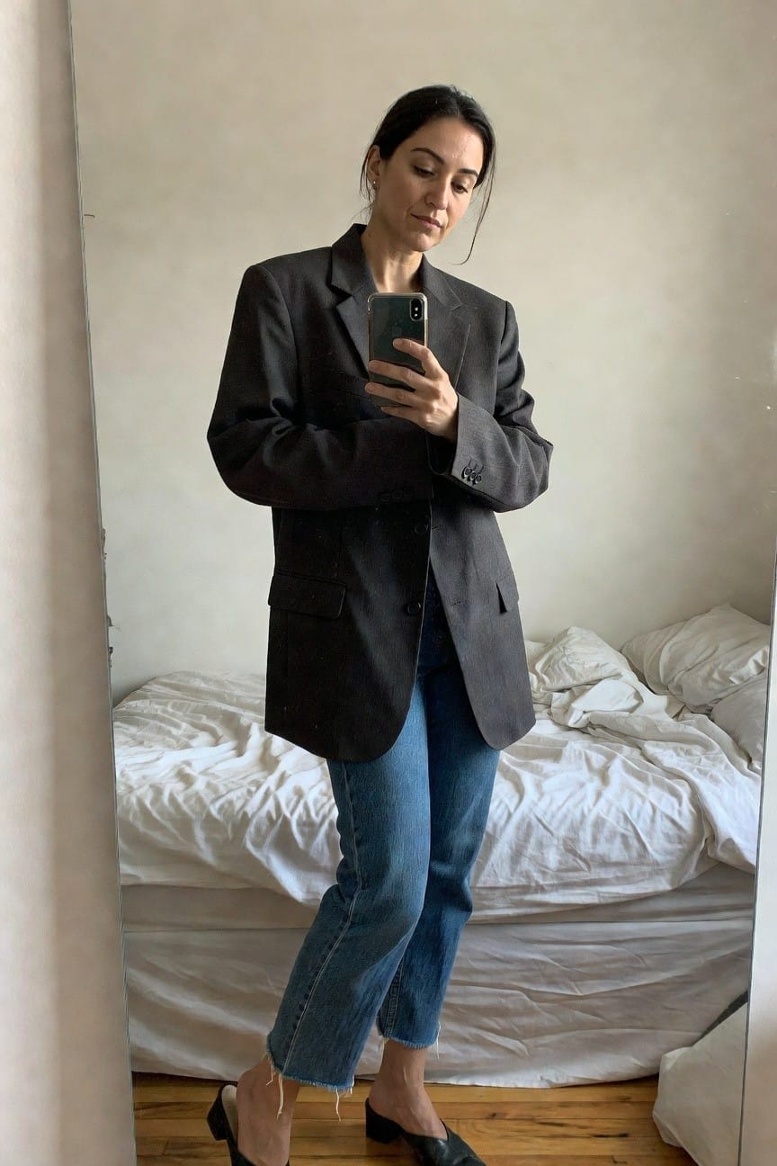 Woman taking mirror selfie in charcoal wool blazer over light blue straight-leg cropped jeans and black platform mules, gold hoop earrings, bedroom with bed in background