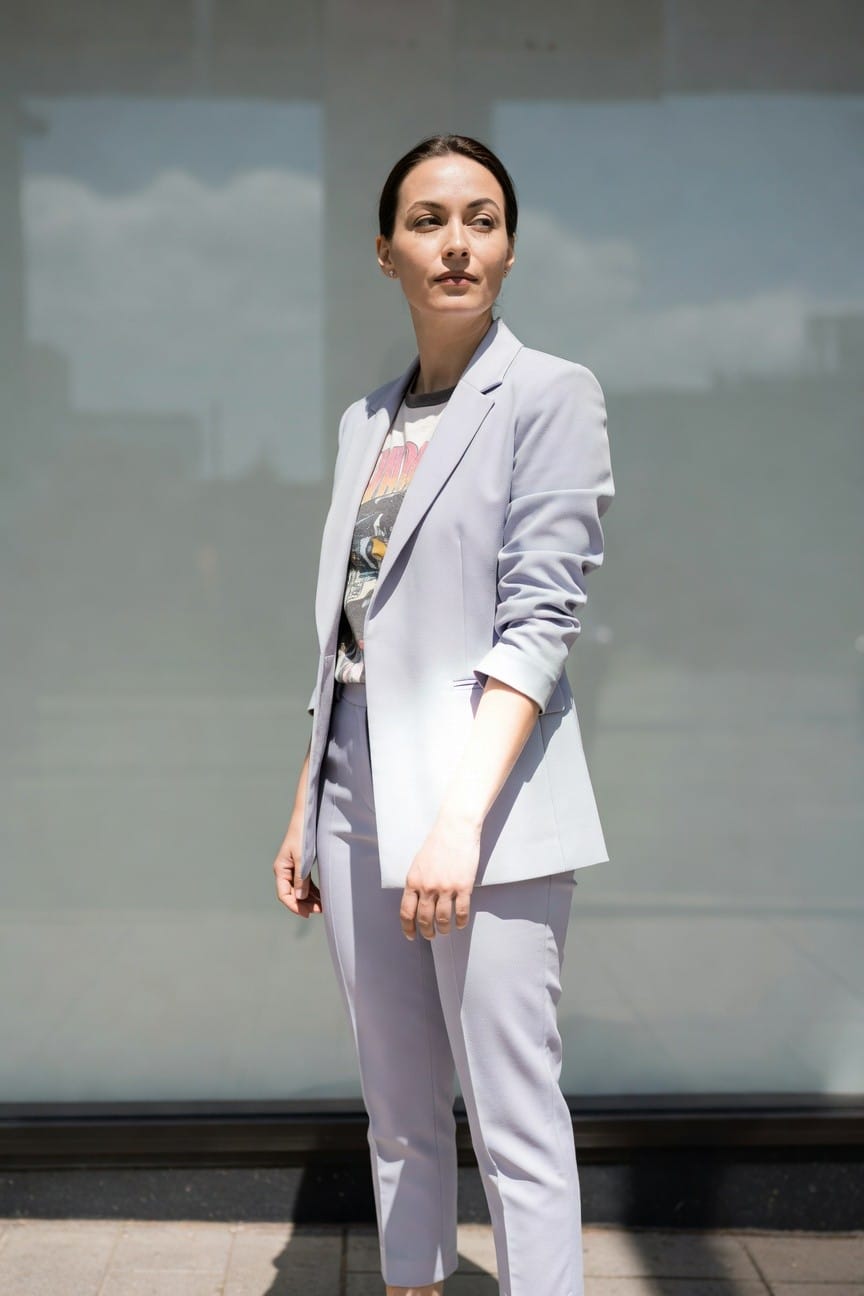 Woman standing confidently in a light gray tailored blazer and slim cropped trousers over a graphic band t-shirt with car print, rolled jacket sleeves, nude heels, and simple earrings against a frosted glass background