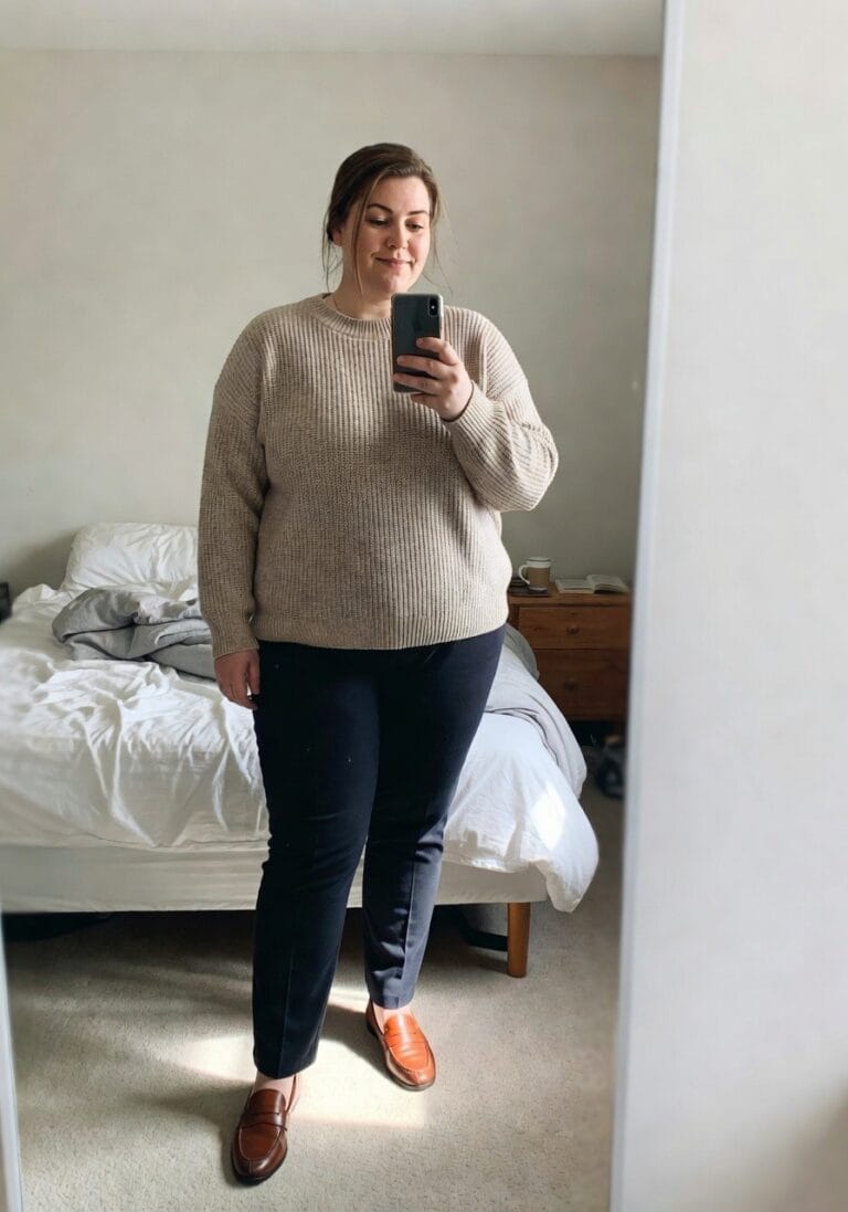 Plus-size woman in mirror selfie wearing an oversized beige knit crewneck sweater, navy straight-leg trousers, and brown loafers, standing confidently in a bedroom with unmade bed in background