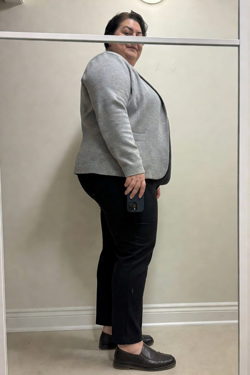 Side profile of a plus-size woman in a light gray knit blazer with open front, black straight-leg pants, and brown leather loafers, standing in a fitting room mirror holding her phone