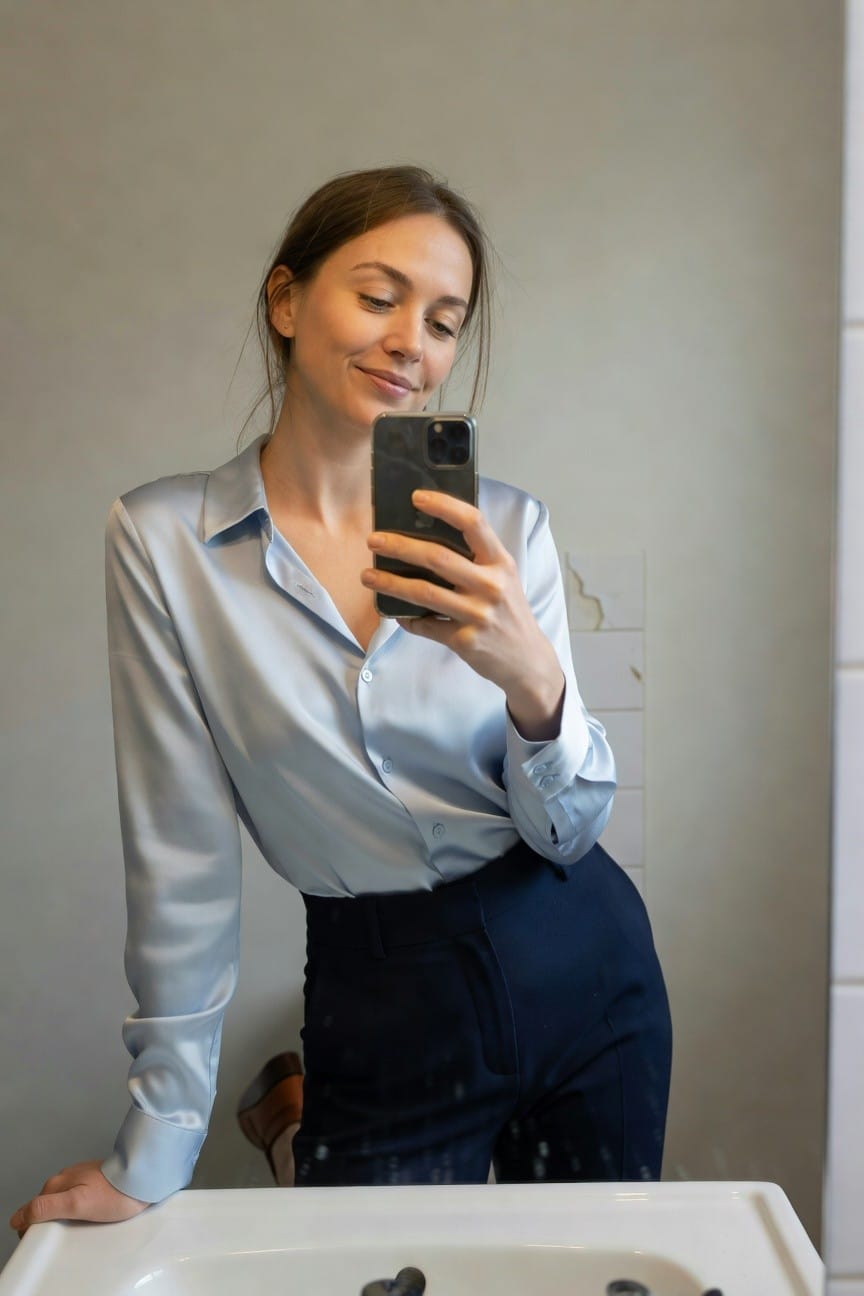 Woman taking a mirror selfie in a bathroom, wearing a light blue satin long-sleeve button-up blouse tucked into high-waisted navy wide-leg trousers, with brown leather heeled ankle boots, one hand on the sink.