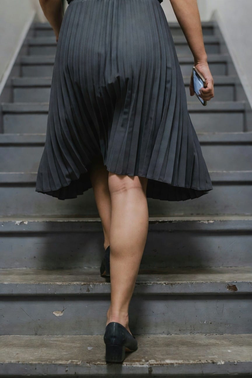 Rear view of woman ascending concrete stairs in a dark gray pleated midi skirt, sleeveless black top, black pumps, holding a smartphone in her right hand