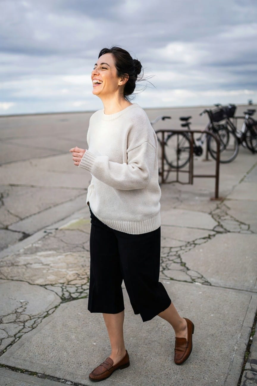 Side view of woman laughing in oversized cream knit sweater, black wide cropped pants, and brown loafers, standing on cracked pavement near bike racks