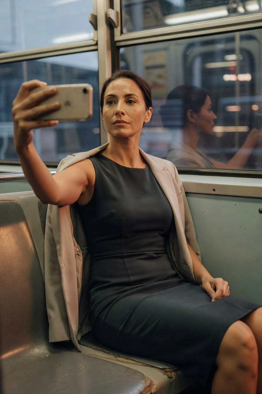 Woman taking a selfie on a train seat, wearing a sleeveless black sheath dress with a beige blazer draped over her shoulders, legs crossed, iPhone in hand
