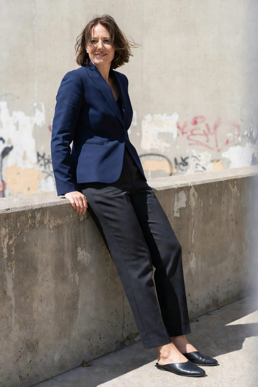 Woman in fitted navy blue blazer and matching slim trousers with black leather mules, leaning casually against a concrete wall