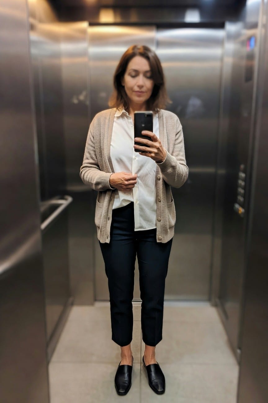 Woman in elevator taking selfie wearing open beige knit cardigan over white collared blouse, navy ankle-length cropped trousers, and black leather mule loafers, neutral polished office outfit