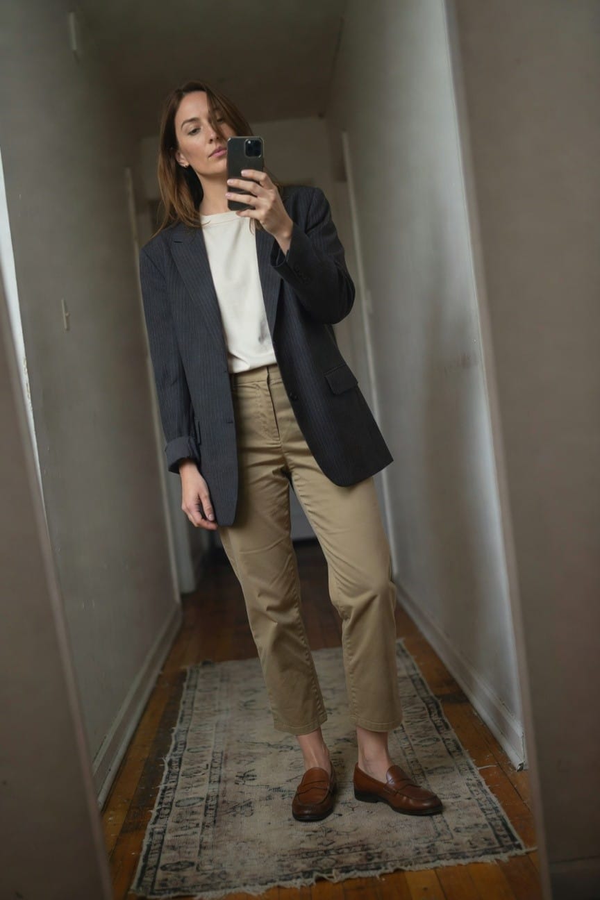 Woman taking mirror selfie in hallway wearing dark gray pinstripe blazer over white crewneck tee, cropped khaki pants, and brown penny loafers, sleeves rolled up on blazer