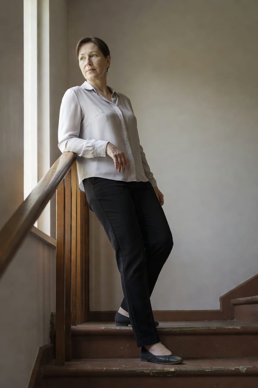 Mature woman in a white long-sleeve blouse, black slim pants, and black ballet flats, leaning casually on a wooden staircase railing indoors