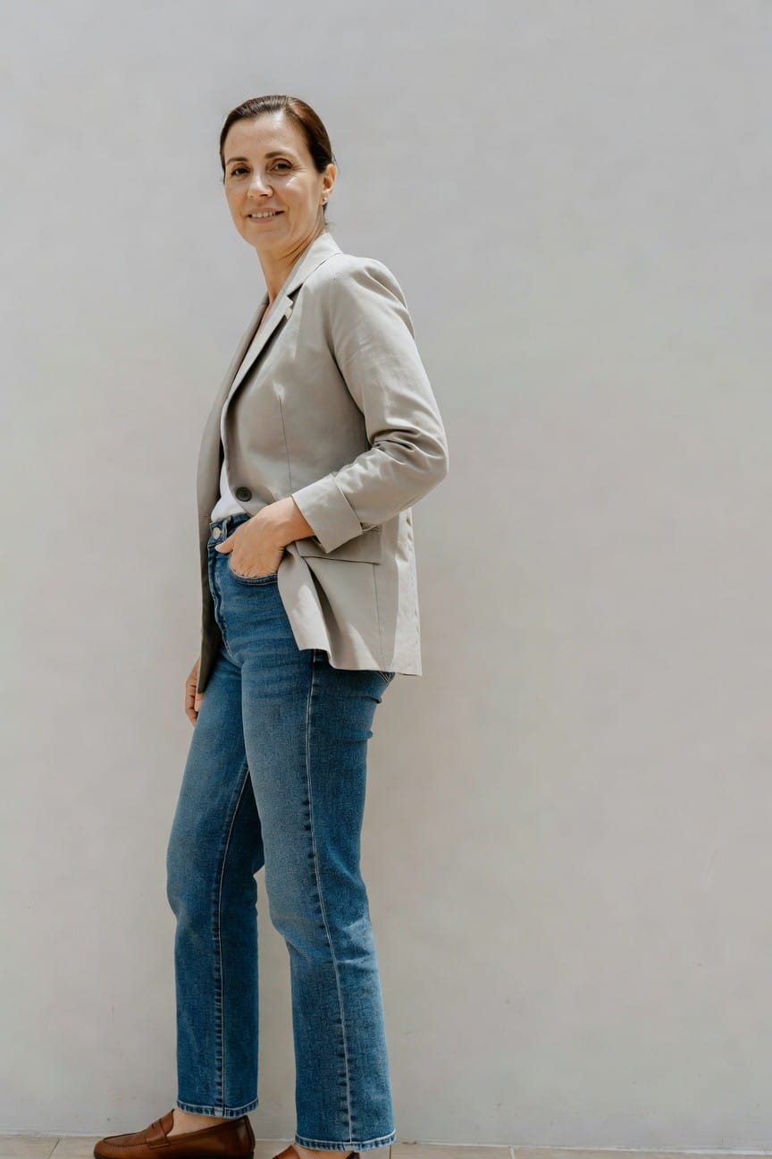 Side view of woman in light gray blazer over white top, high-waisted straight blue jeans, and brown loafers, hand in pocket, standing confidently