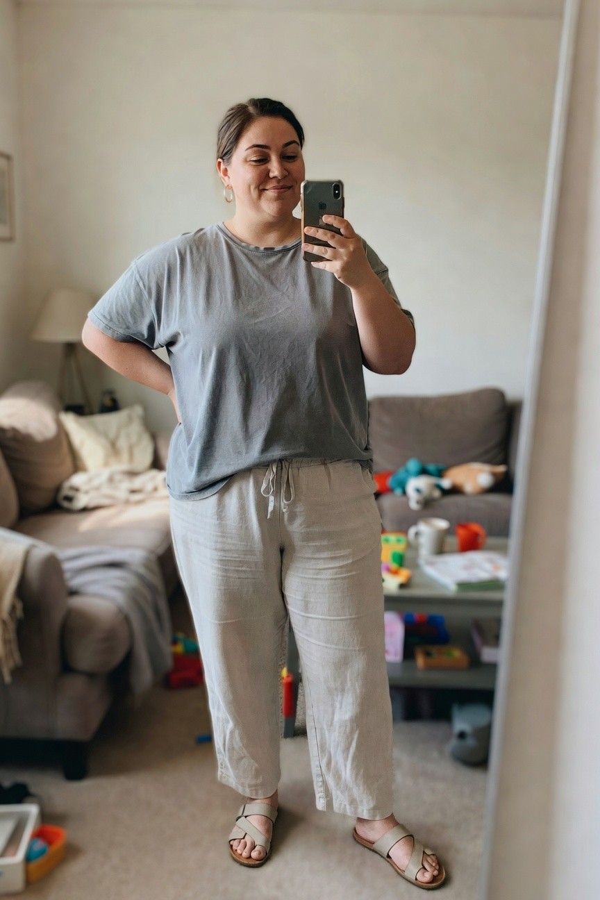 Woman in gray tee and beige pants taking a mirror selfie in a living room.