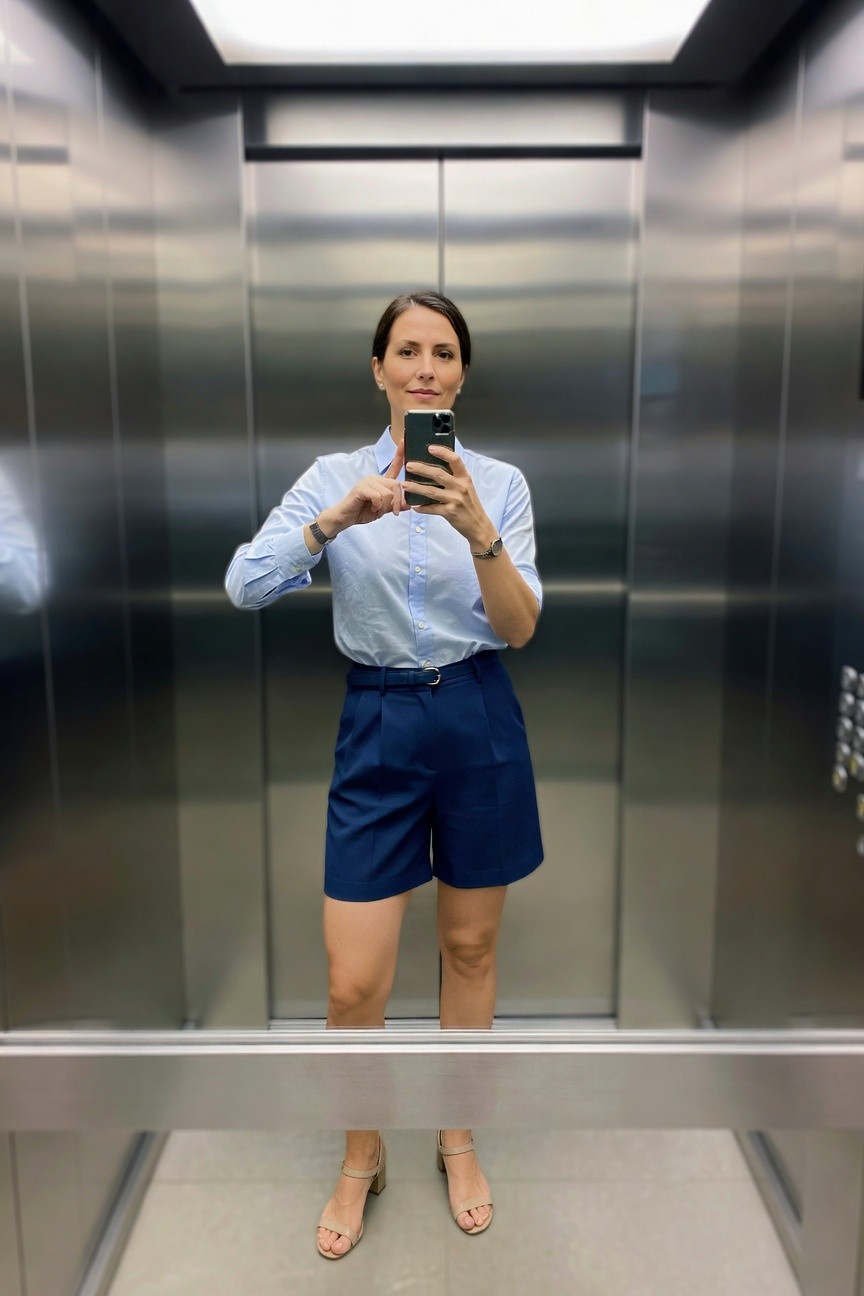 Woman wearing a light blue button-up shirt tucked into navy tailored shorts with a belt and beige sandals while taking a mirror selfie in an elevator.