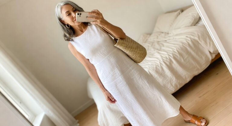Woman over 60 wearing a white sleeveless linen midi dress with brown leather cutout sandals and a woven straw shoulder bag while holding a smartphone.